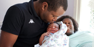 A father kisses his child in the hospital after birth, while his wife, who is lying down, smiles.