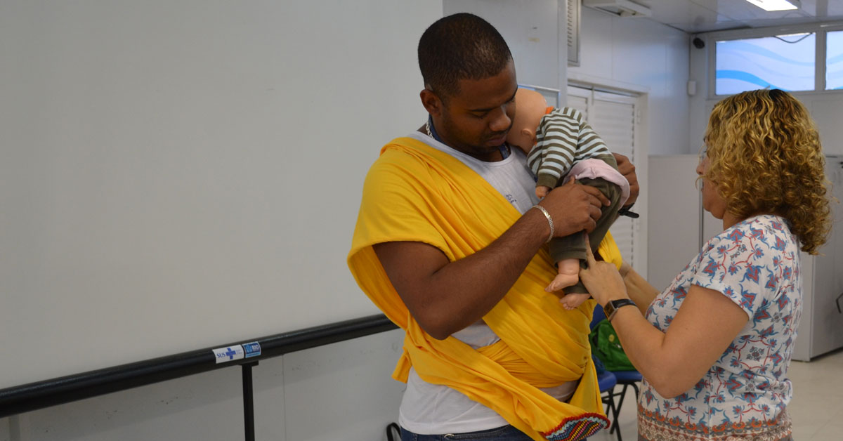 A father learns how to carry a baby in a sling, using a doll, in a MenCare+ workshop in Brazil.