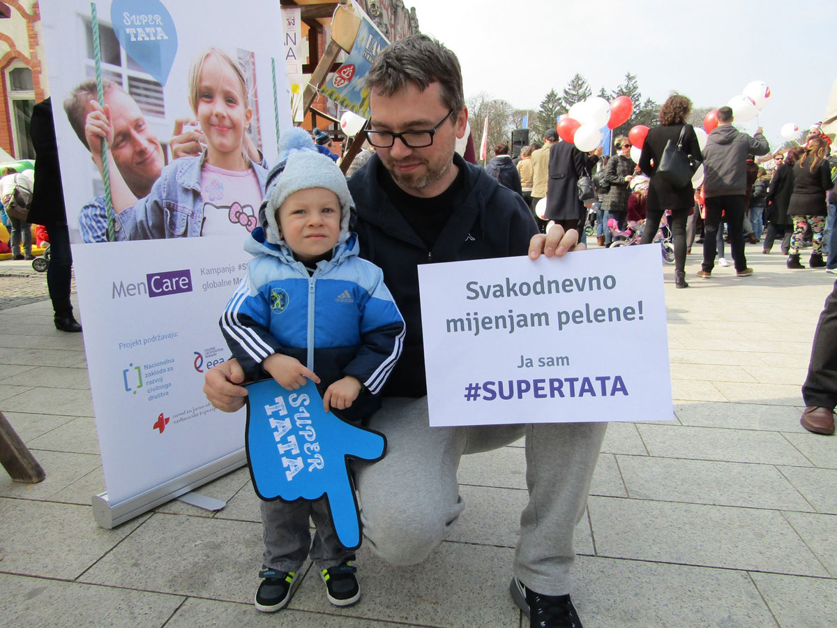 A father holds a sign that reads: “I change diapers daily.”