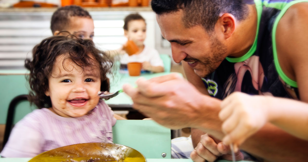 A smiling father feeds his laughing daughter.